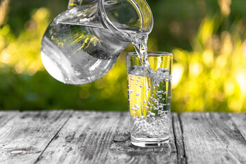 Clean water pours into a glass from a glass jug on a background of greenery in the fresh air....