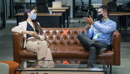 Male employee sat with a female colleague on the sofa wearing a mask and spaced social space.New...