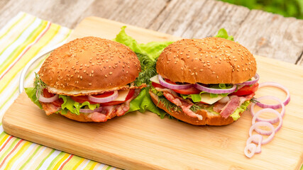 Picnic sandwiches with lettuce, bacon, onion rings, tomatoes, egg, cheese and sesame bun on a background of greenery in the fresh air.