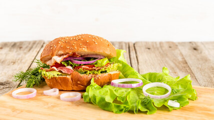 Sandwich with lettuce, bacon, onion rings, tomatoes, egg, cheese and sesame bun on a wooden background.