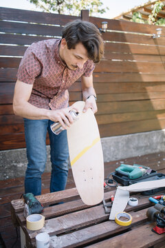 Focused human holding skateboard while painting it outdoor. Brunette man standing in yard and holding skateboard while using spray paint.