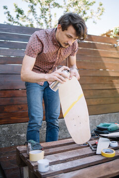 Focused human holding skateboard while painting it outdoor. Brunette man standing in yard and holding skateboard while using spray paint.