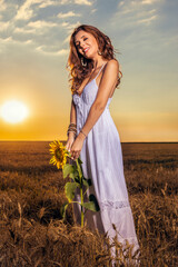 Obraz premium Beautiful woman in white dress in a wheat field with a sunflower flower at sunset.