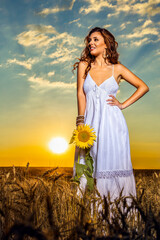 Obraz premium Beautiful woman in white dress in a wheat field with a sunflower flower at sunset.