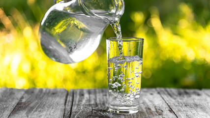 A glass glass is filled with water from a jug on a background of green grass in the fresh air. A...