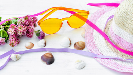 Obraz na płótnie Canvas Yellow rimless glasses between a straw hat and pink flowers on a white background among seashells. The concept of summer vacation.
