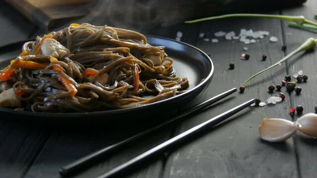 Freshly boiled steam asian buckwheat noodles soba with vegetables onion pods, carrots, paprika and pieces of chicken, on a stylish black plate and Chinese chopsticks.
