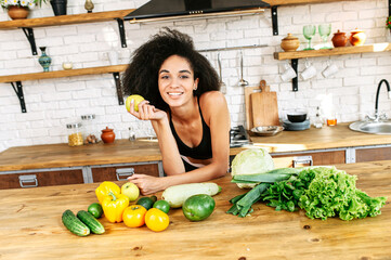 Eat healthy food. Fresh vegetables and greenery lay on the table in the kitchen, young beautiful african-american woman holds fresh green apple in hand and looks at camera with a smiles