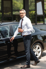 Smiling businessman looking at camera while holding paper cup near auto on urban street