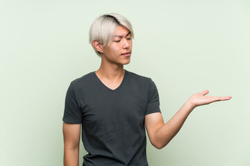 Young asian man over isolated green background holding copyspace with doubts