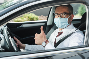 Fototapeta na wymiar Selective focus of businessman in medical mask showing thumb up while driving auto