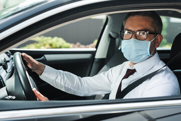 Selective focus of businessman in eyeglasses and medical mask looking at camera while driving car