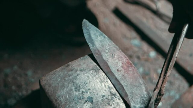 Steel knife blade held by metal tongs hammered on anvil