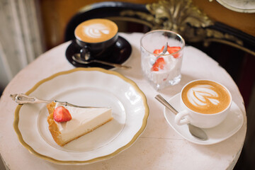 Cheesecake, ice cream with strawberries and cups of cappuccino  on marble table in beautiful interior.