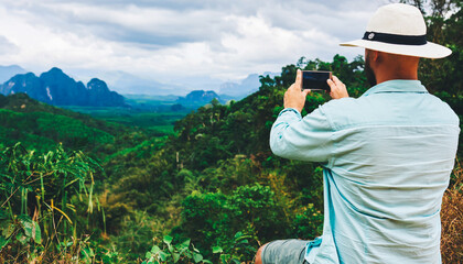 Fototapeta na wymiar Back view of a man wanderer is shooting video on smart phone, while is sitting on a mountain against subtropical forest. Young hipster guy is taking photo on cell telephone of a beautiful jungle view
