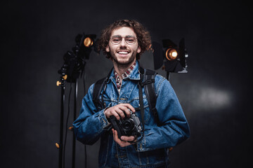 Portrait of a handsome curly guy in glasses wearing denim jacket holds a digital camera, smiling and looking on camera. Dark photo studio with lighting equipment in the background