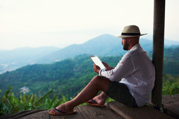 Young man wanderer is reading electronic book on touch pad, while is sitting against fantastic...