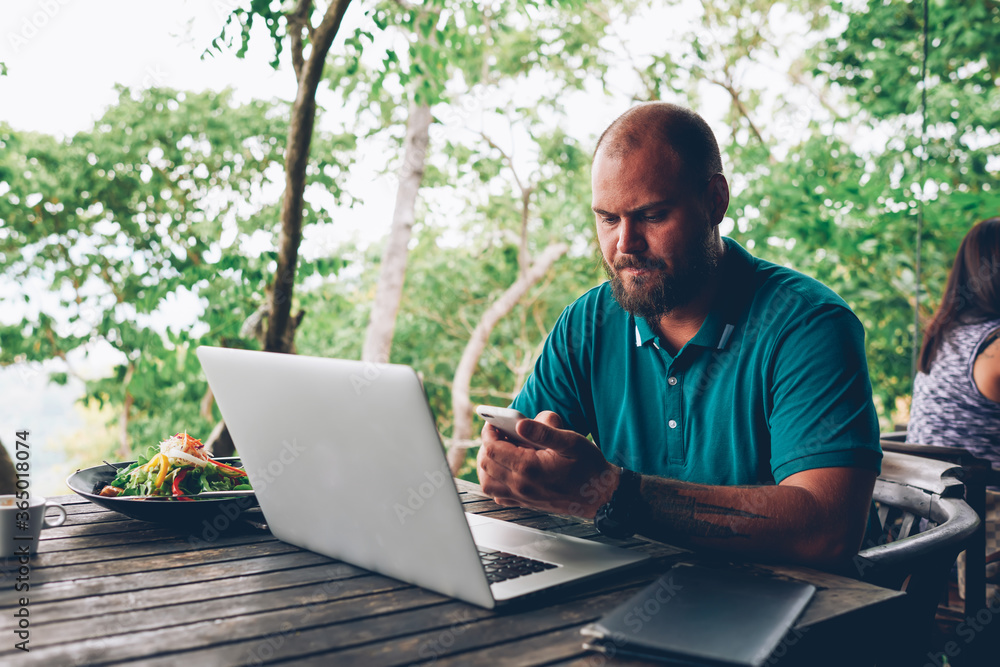 Poster serious hipster guy making payment for salad on lunch using banking application on smartphone, man f