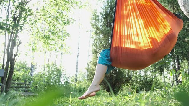 Woman reading e-book or using tablet while sitting in hammock chair on nature on sunset. Cottagecore, slow life in countryside, relax outdoor, summer vacation concept.