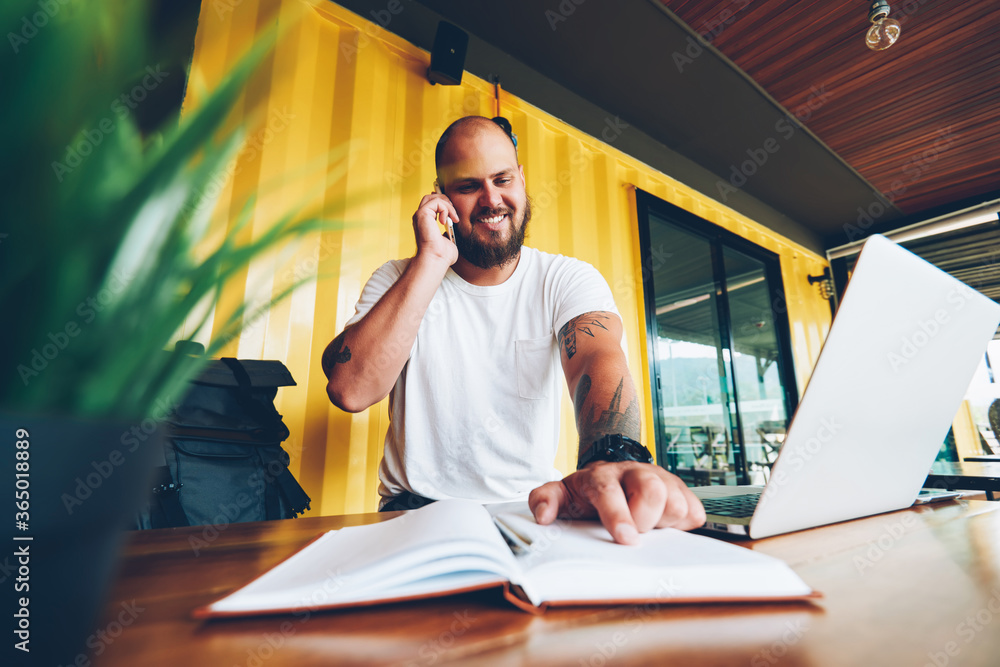 Poster cheerful hipster guy pointing on notes in notebook share information during mobile phone talk. happy