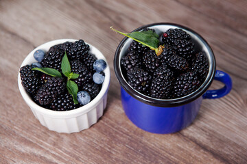 Fototapeta na wymiar Fresh berries of blackberries, blueberries, mint leaves, in white bowl and blue mug on wooden background.