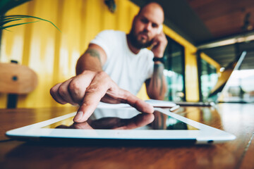 Portrait of serious bearded man looking at camera making phone call and touching tablet for zoom, handsome man enter security code on portable pc while having cellphone conversation in office