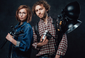 The team of two young photographers holds a digital camera and lighting equipment posing in studio
