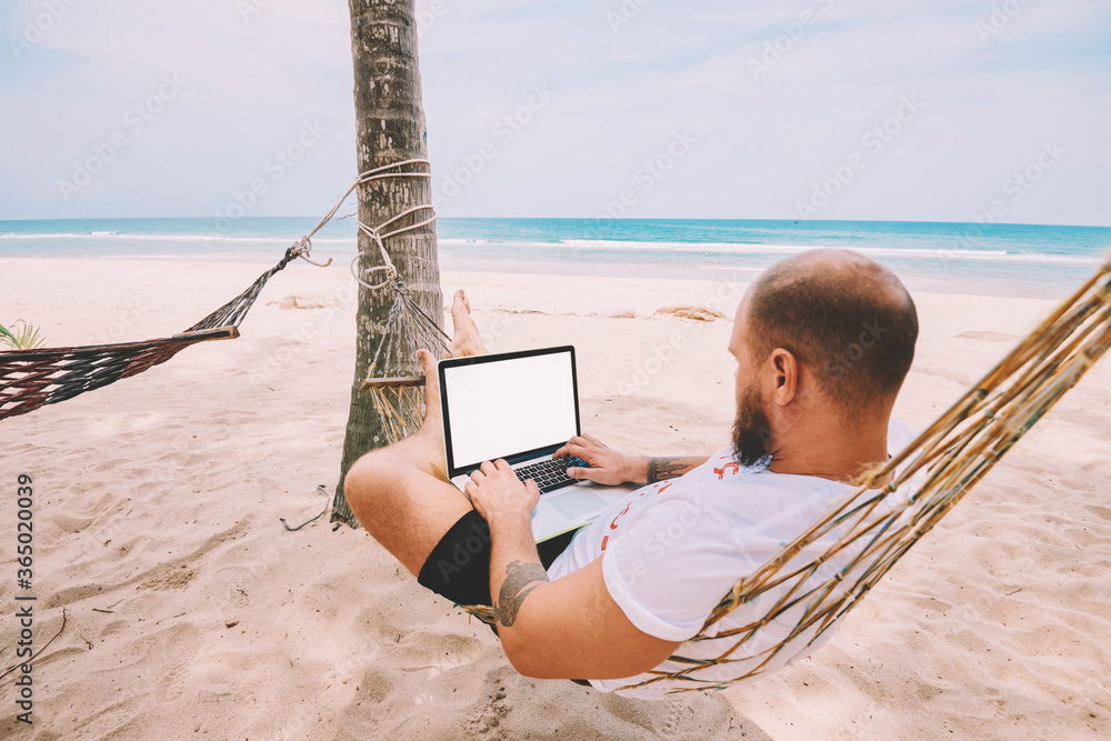 Poster young man resting on hammock while doing work freelance remotely at modern laptop computer with mock