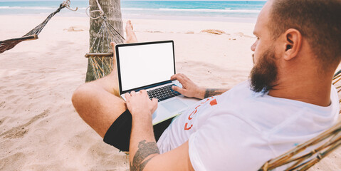 Male businessman tourist typing text on keyboard of computer with blank screen for your internet website rest on tropic island. Young man freelancer work at laptop during distance job lying on hammock