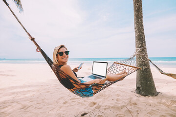 Happy young woman freelancer entrepreneur in sunglasses smiling at camera while lying on hammock with modern laptop computer and working remotely on seashore of tropical island during summer vacations