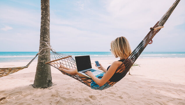 Young woman tourist reading notification on smartphone in roaming while lying on hammock and working remote at laptop computer with mock up area for your content during summer vacation on ocean beach