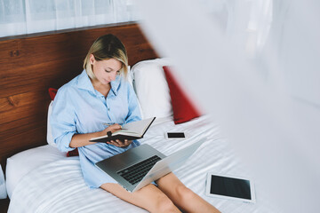 Concentrated young female blogger writing article for publication in notebook lying in comfortable bad, young thoughtful woman noting information working remotely from hotel room on vacation