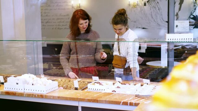 positive young women with cross body handbags choose delicious fresh cakes and candies standing near glass display in modern confectionery shop