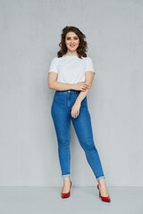 Positive girl in jeans, t-shirt and stilettos posing near the wall