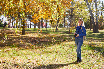 Obraz premium Attractive beautiful young girl holding modern mirror camera in autumn park. Traveller walking in place with yellow leaves on tree at background