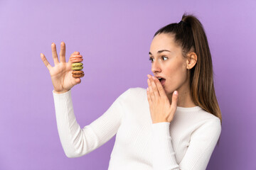 Young brunette woman over isolated purple background holding colorful French macarons and with surprise expression