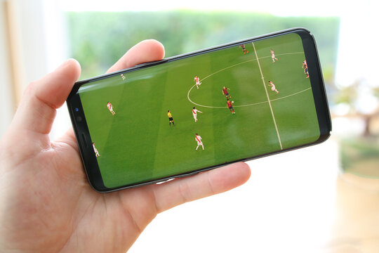 A male hand holding a mobile phone which displays a soccer match on the touch screen. The big green screen is in contrast with the overexposed background. An image on an interior background.
