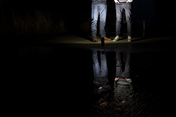 Two anonymus young men standing in front of a pool of water, during night time. Their reflection is...