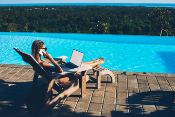 Young sexy woman in sunglasses talking on smartphone while doing distance job at laptop with blank...