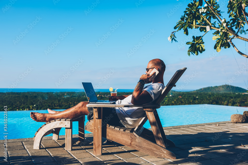 Poster young businessman resting on sunbed enjoying summertime in resort spa near swimming pool while worki