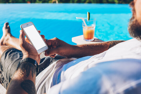 Cropped image of man lying near swimming pool with cocktail and chatting online via high speed internet connected to modern smartphone.Male installing app on mobile phone enjoying summer vacation