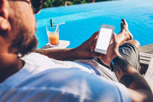 Cropped image of male blogger holding modern smartphone with blank copy space screen for your advertising information content in hands resting on sunbed with cocktail near blue swimming pool
