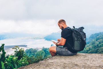 Bearded hiker with backpack sitting on top of hill with amazing scenery of landscapes and mountains...