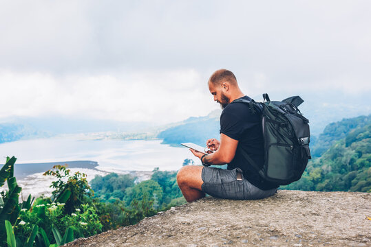 Bearded hiker with backpack sitting on top of hill with amazing scenery of landscapes and mountains with green vegetation washed by sea while drawing pictures of nature with on modern tablet