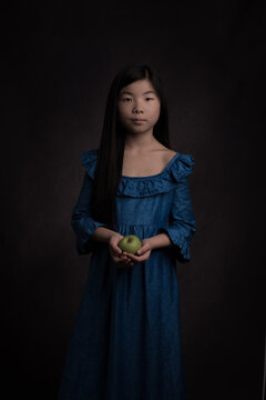 Girl in vintage classic blue dress and long black hair holding a green apple in a renaissance painterly studio portrait