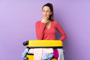 Traveler woman with a suitcase full of clothes over isolated purple background thinking an idea