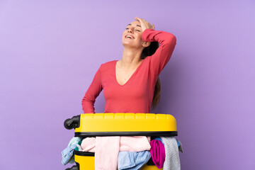 Traveler woman with a suitcase full of clothes over isolated purple background laughing