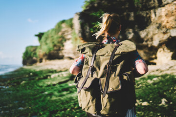 Young blonde woman tourist with stylish backpack travelling during summer vacation.Hipster traveller with rucksack walking in windy weather on green island with high hills on weekend