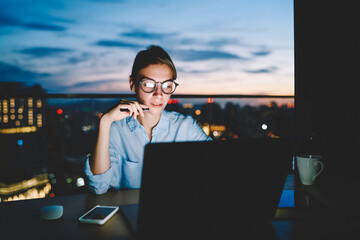Pensive female freelancer working on publication for share on web page sitting with laptop at...