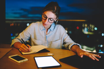 Concentrated businesswoman staying overtime writing plan for startup in notepad, clever  female student preparing for examination making notes and using modern technologies at working place at home